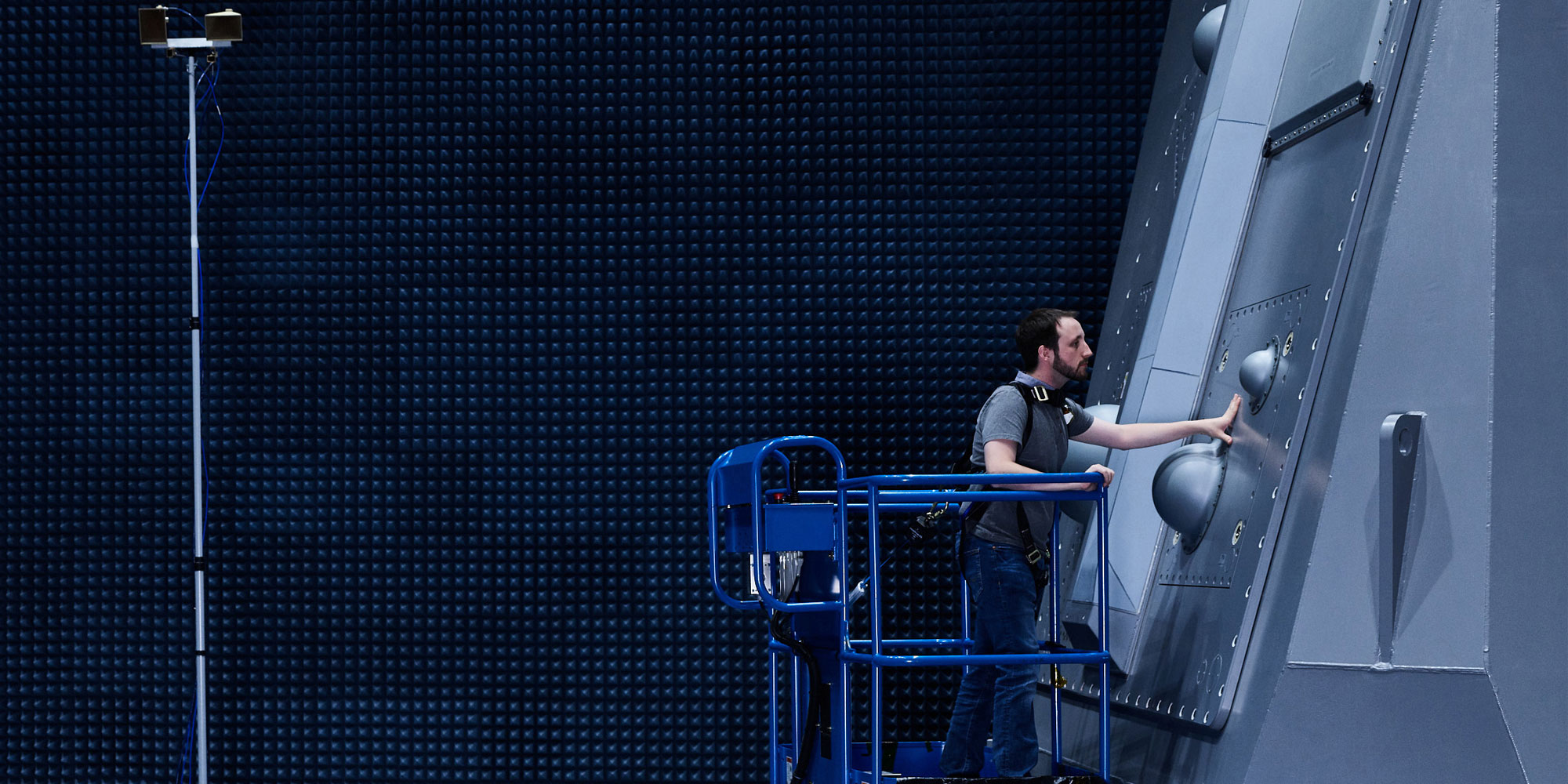 Technician inspecting naval radar system in anechoic chamber using blue safety lift platform indoors.
