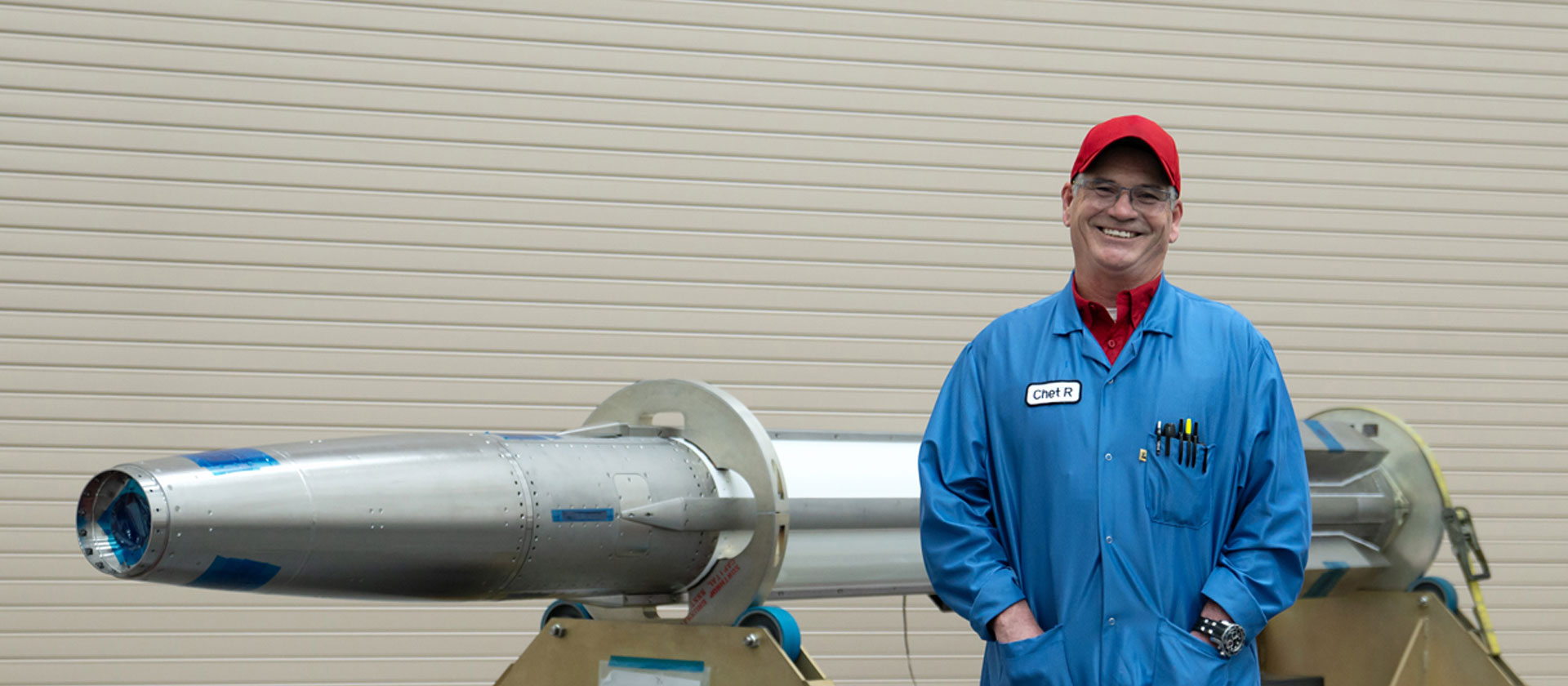Man wearing blue jacket and red hat standing in front of a rocket.
