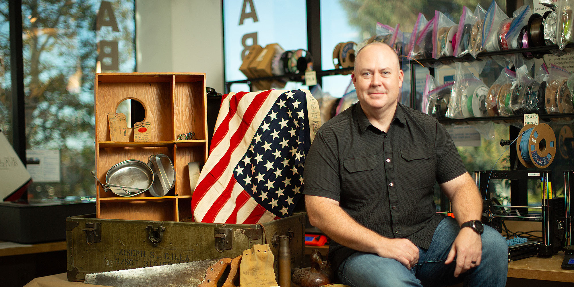 Man sits on World War II footlocker with memorabilia. 