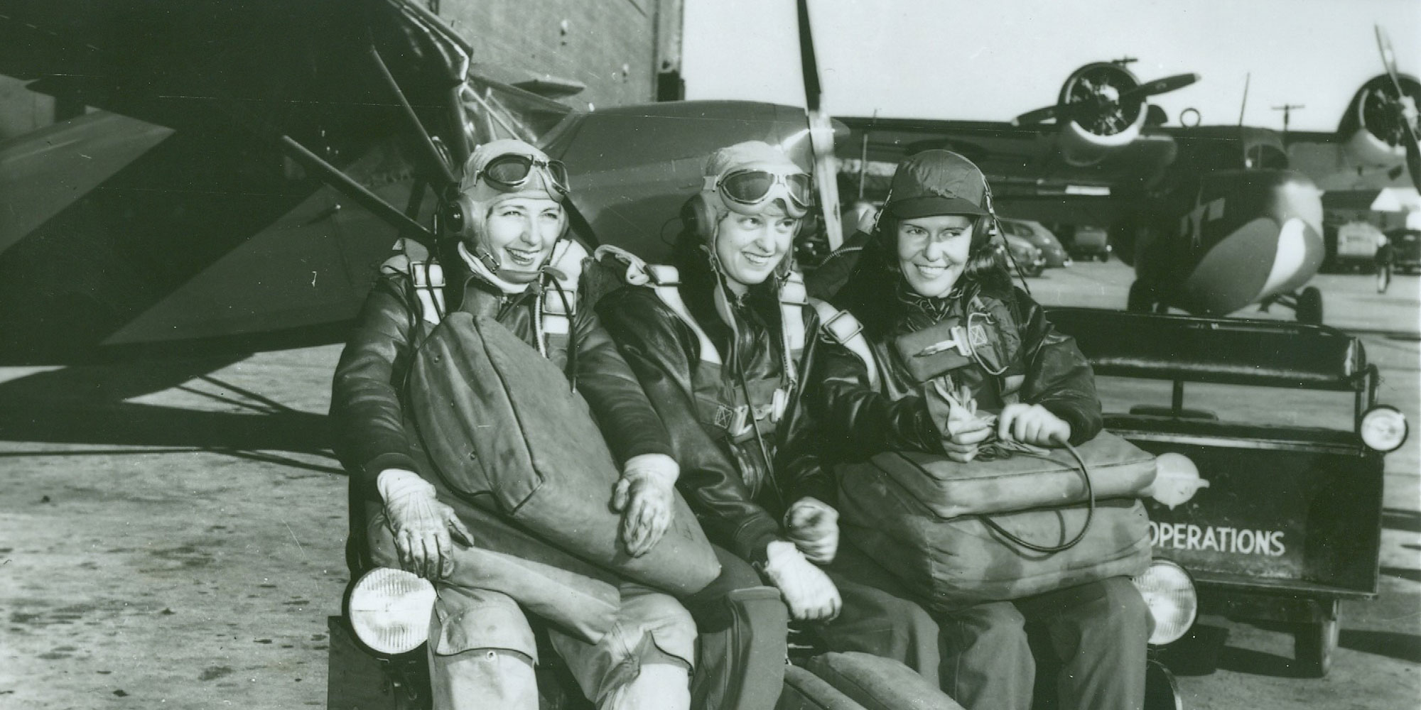 Black and white image of three women in flight suits on a runway, sitting and smiling.