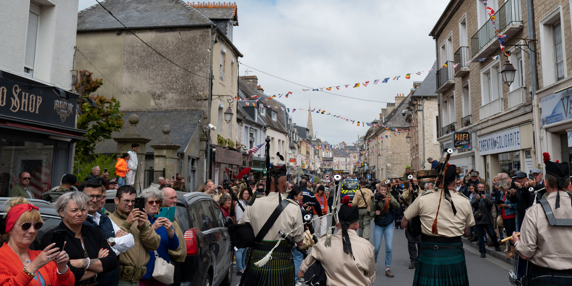 Crowds of people watch a parade featuring bagpipers in a French town with flags flying