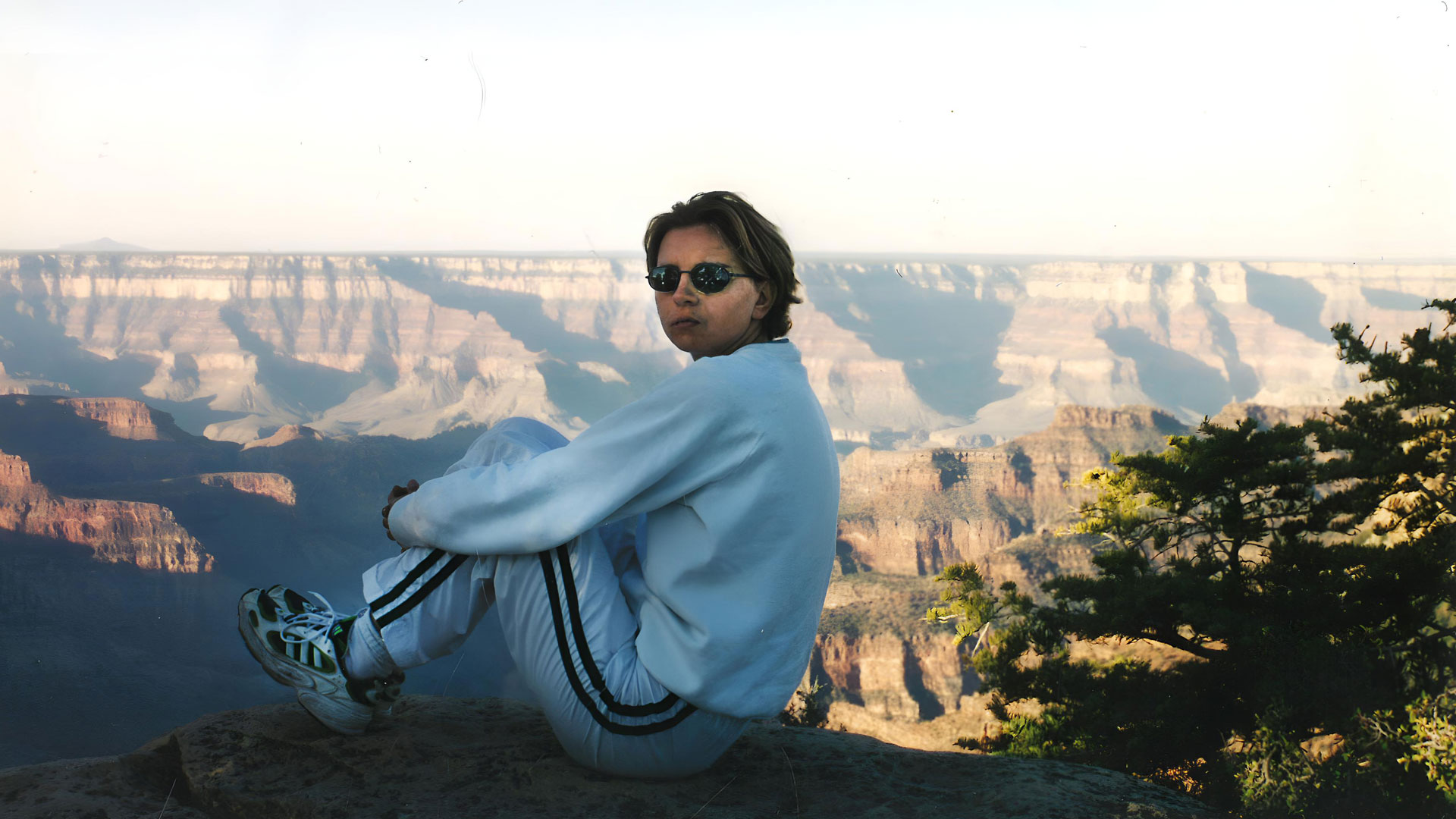 A woman in a blue tracksuit and sunglasses sits in front of the Grand Canyon.
