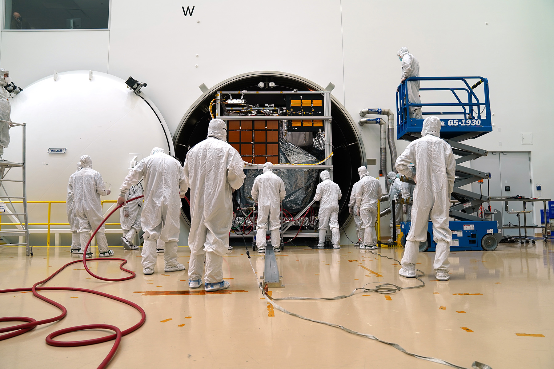 Engineers facing forward in full covering lab coats are preparing a satellite for testing