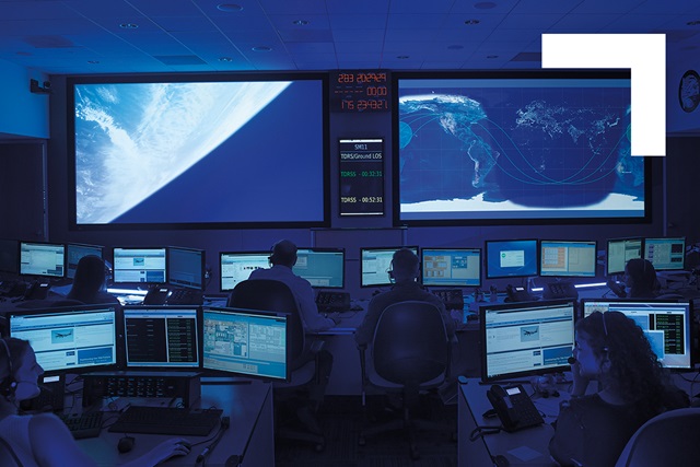 Two large projection screen monitors in data center with several workstations with dual monitors and various employees working