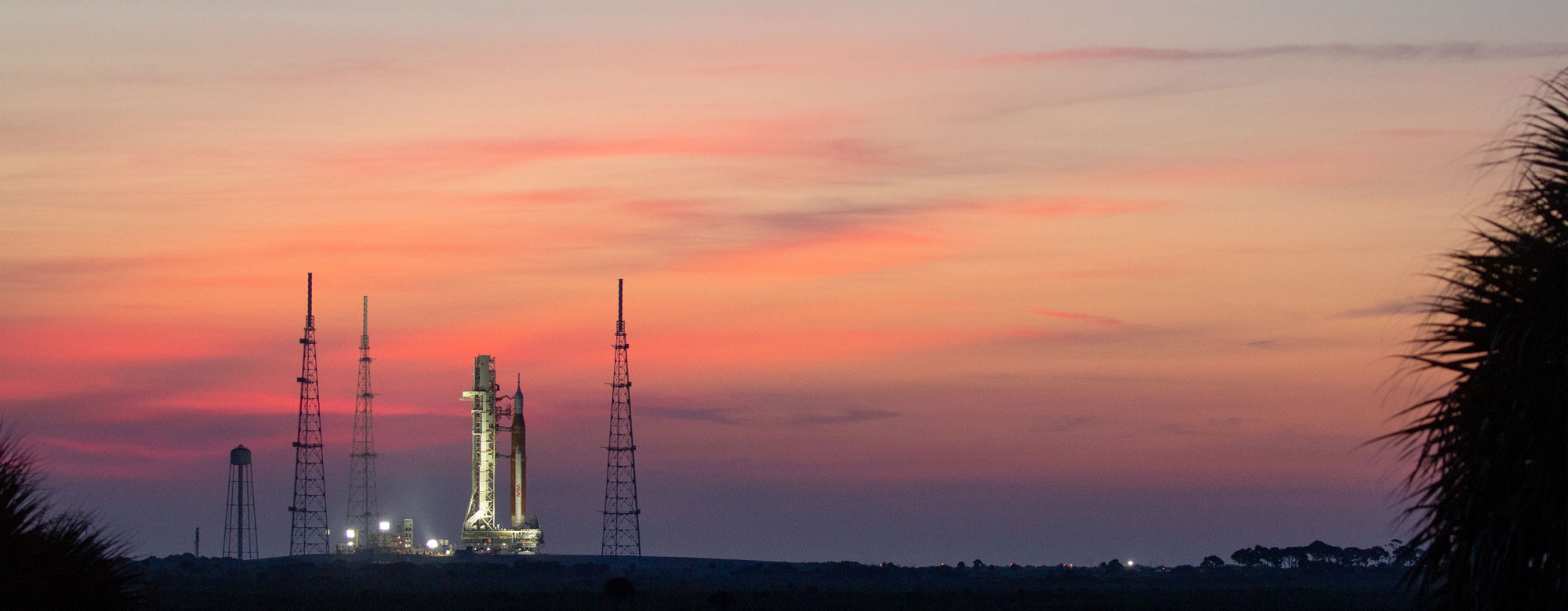 artemis-women-article View of Artemis I before launch with a view of water and a pink and purple sky.